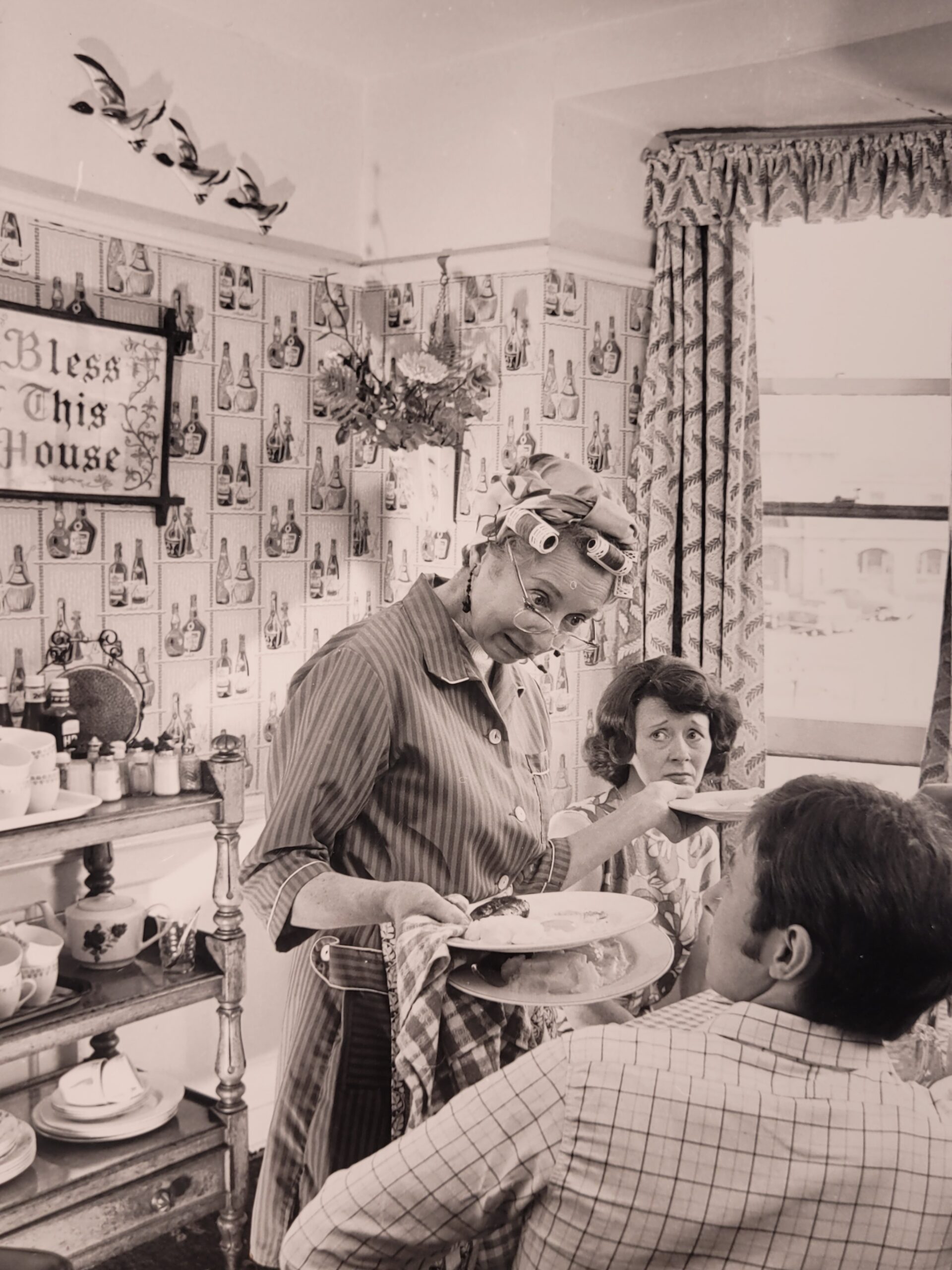 Family meal, housewife serving bangers and mash in curlers stereotypical british sixties household, by Michael Joseph
