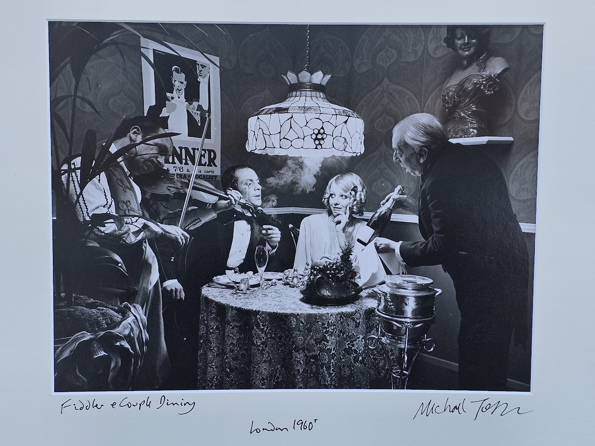 Fiddler and couple dining with art deco poster behind, test print for UK Shell & BP magazine advert by Michael Joseph