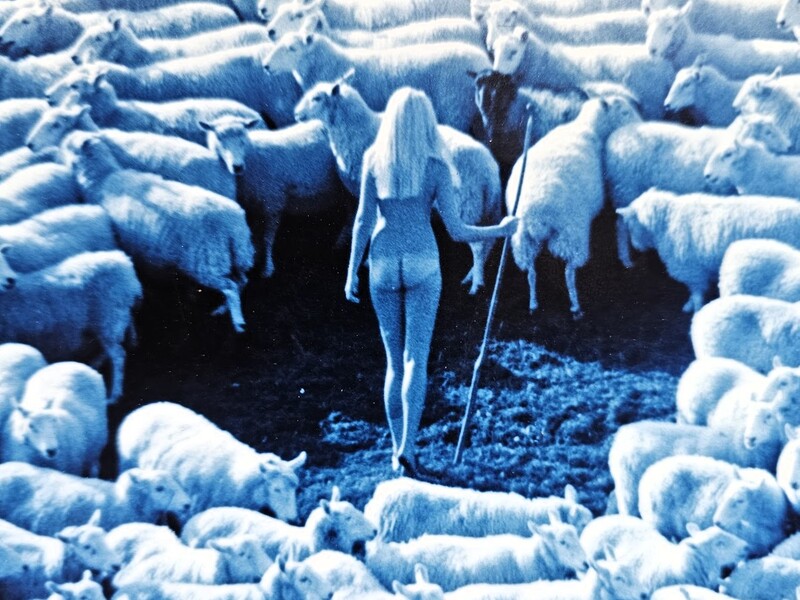 A blue-toned image of a nude woman holding a staff surrounded by sheep