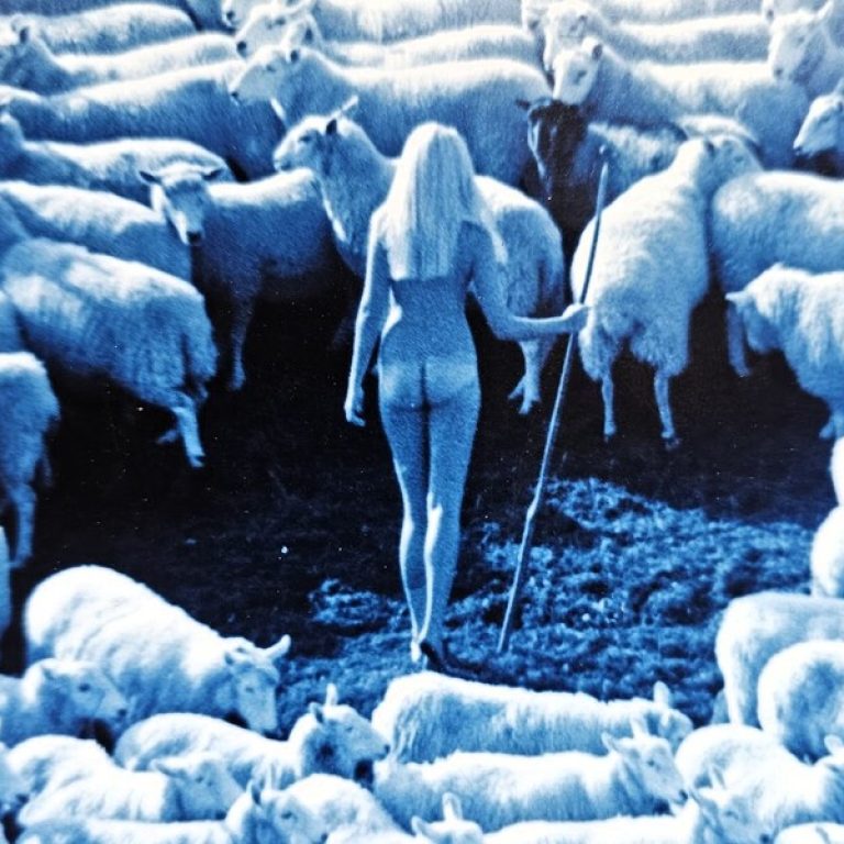 A blue-toned image of a nude woman holding a staff surrounded by sheep